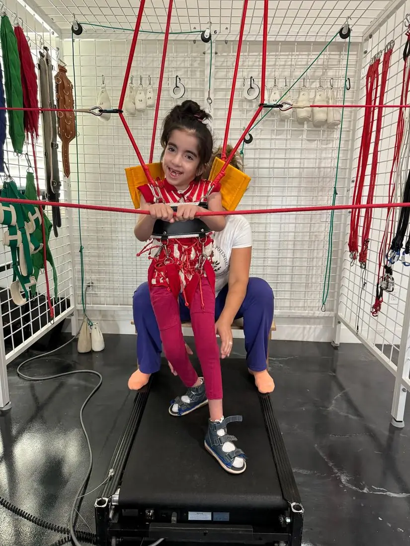 A smiling girl walks on a treadmill inside the SpiderCage with a therapist's support